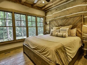 A large bed with a wooden headboard in a room with large windows and a view of trees.