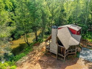 Eine Hütte mit geneigtem Dach, Schornstein und einer Veranda, umgeben von dichtem Wald und einem Bach.