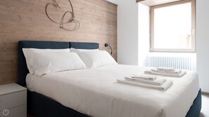 A neatly made bed with white linens and folded towels, a wooden headboard, and a window with blinds. - Roma 38 - Two Bedroom Apartment, Sleeps 6 (Bormio)