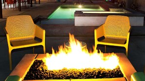 Et bålsted med to stole på hver side, en pool i baggrunden og en terrasse af beton.