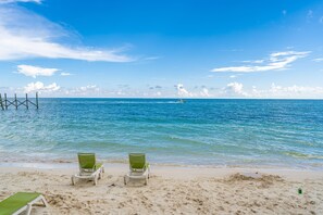 Een strand met twee groene ligstoelen gericht op de oceaan, een steiger die in het water uitloopt, en een helderblauwe lucht.