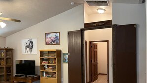 Sala de estar com televisão, estantes de madeira e uma porta que dá para outro quarto.