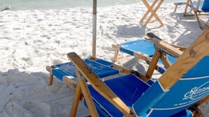 Escena de playa con una sombrilla azul, sillas de madera para tomar el sol y vista al océano.