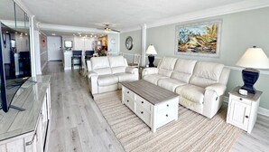 Un salón espacioso con sofá, mesa de centro y televisor. Se observa una cocina al fondo con armarios blancos y un comedor con sillas.