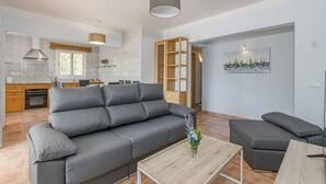 Een moderne woonkamer met een grijze bank, een houten salontafel en een grijze fauteuil. Achtergrond toont een keuken met houten kastjes en een raam.