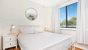 A bedroom with a bed, two bedside tables, a round mirror, and a window with curtains. - Scenic Shack - 4 Scenic Way (CARRICKALINGA)