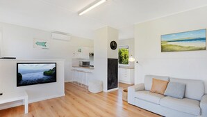 A modern living room with a flat-screen TV, a sofa, and a kitchen area with a dining table. - Scenic Shack - 4 Scenic Way (CARRICKALINGA)