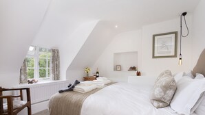 A bedroom with a large bed, a window with curtains, a wooden chair, and a bedside table with a vase of flowers. - Attractive thatched roof property in the idyllic Cotswold village of Ebrington. (Ebrington, near Chipping Campden)