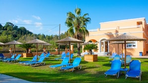 Ein Resort mit mehreren Liegestühlen und Reetdachschirmen, einer Palme und einem Gebäude mit heller Fassade.