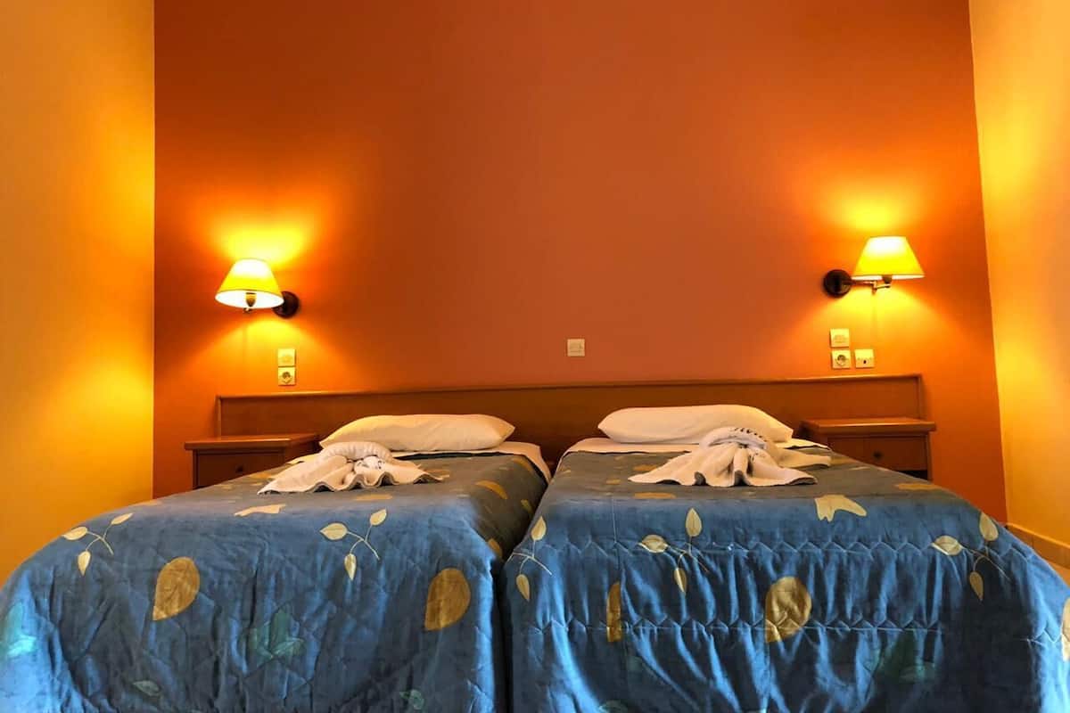 A hotel room with two beds, each with a blue bedspread and white pillows. There are two wall-mounted lamps on either side of the bed.