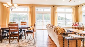 A spacious living room with a dining area, large windows, a ceiling fan, and a view of a forested area. - Kidd Island (CDA) Lakefront Beauty-Amazing views! (Coeur d'Alene)