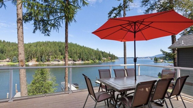 A deck with a table and chairs, overlooking a lake with a forested shore and a boat dock. - Kidd Island (CDA) Lakefront Beauty-Amazing views! (Coeur d'Alene)