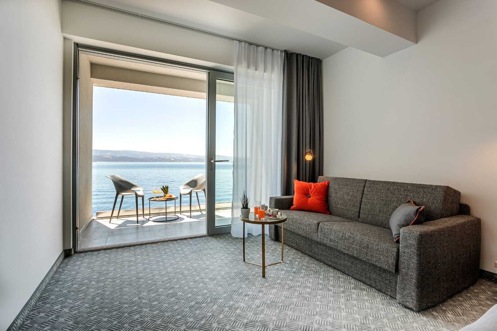 Habitación de hotel moderna con un sofá gris, dos sillas, una mesita y vista al mar a través de un ventanal corredero.