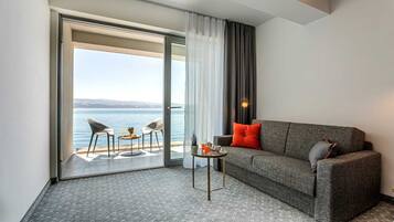 Habitación de hotel moderna con un sofá gris, dos sillas, una mesita y vista al mar a través de un ventanal corredero.