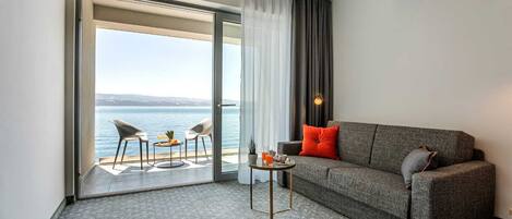 Habitación de hotel moderna con un sofá gris, dos sillas, una mesita y vista al mar a través de un ventanal corredero.