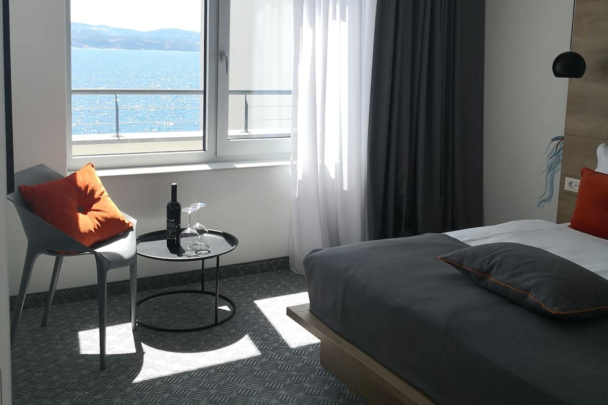 Ein Hotelzimmer mit Bett, Sessel, kleinem Tisch, Flasche und Blick aufs Meer.