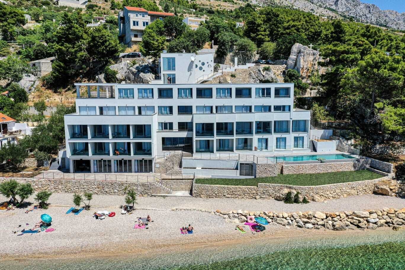 A multi-story hotel with a pool, located on a hillside with a beach in the foreground.