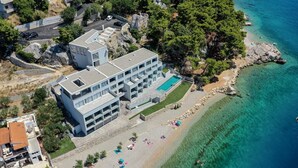 Un edificio moderno de varios pisos con piscina, situado cerca de una playa de aguas turquesas cristalinas y afloramientos rocosos.