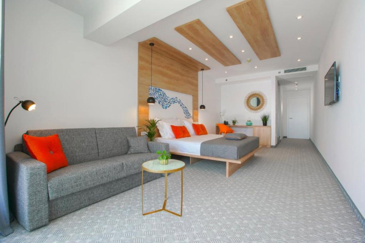 A modern living room with a grey sofa, a small round table, and a bed with orange pillows.