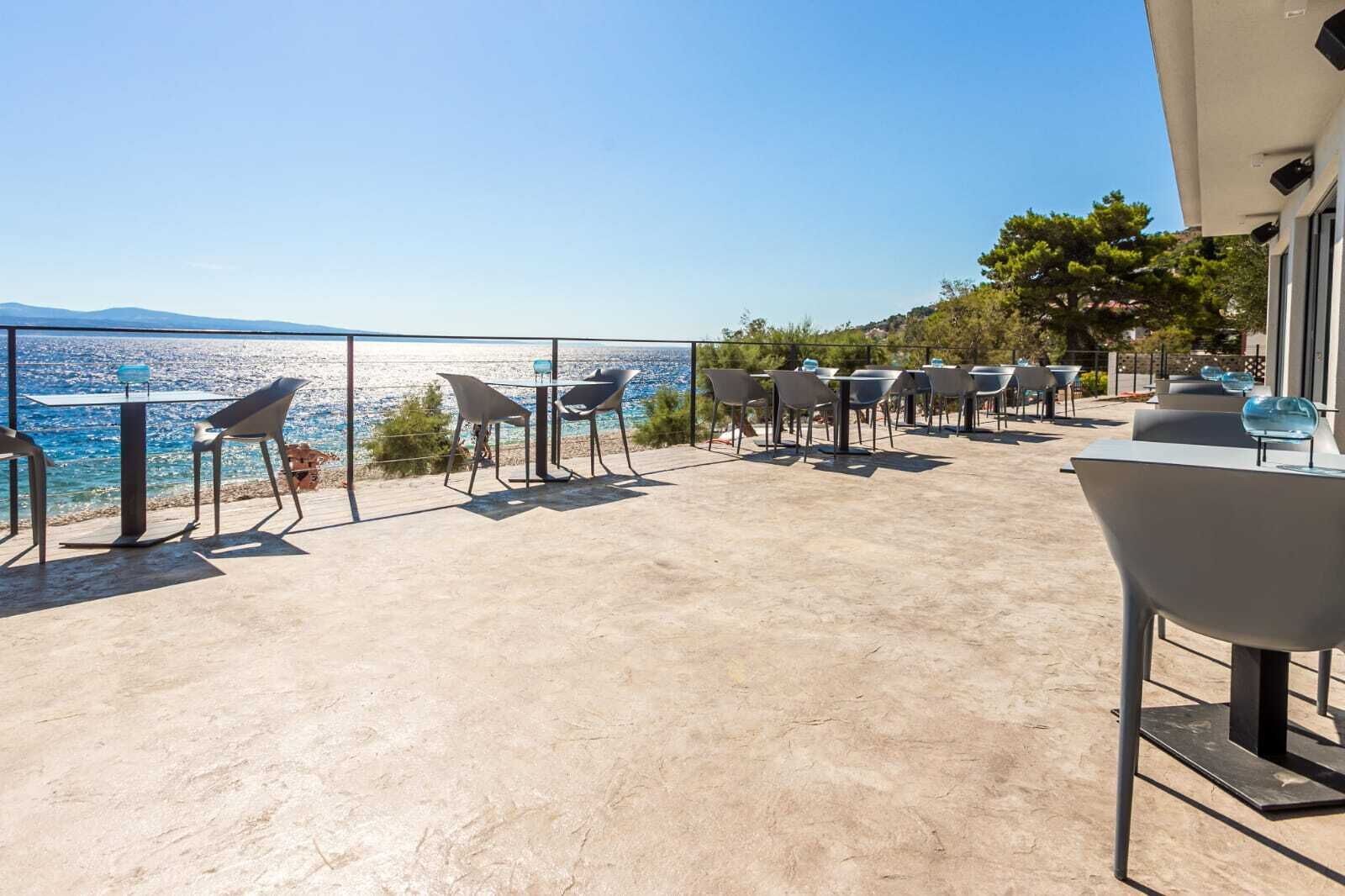 An outdoor terrace with tables and chairs, overlooking a coastal area with a clear view of the sea and distant mountains.