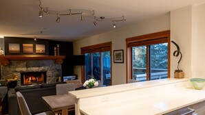 A modern kitchen with a stone fireplace, a dining area with a bench, and large windows offering a view of the outdoors.