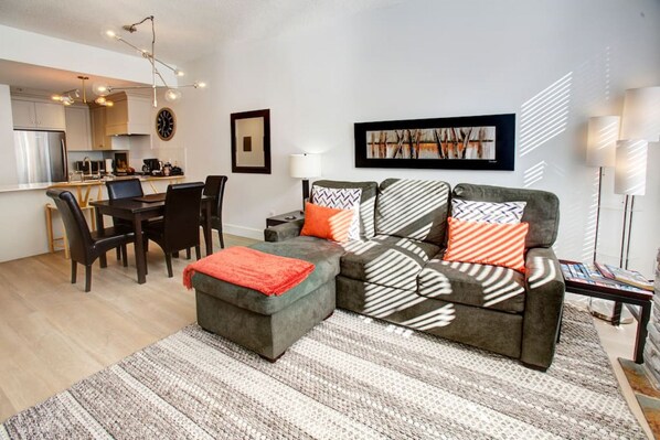 A modern living room with a sectional sofa, a dining table with chairs, a kitchen area, and a large abstract painting on the wall.