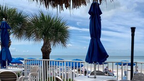 An outdoor seating area with white chairs and blue umbrellas, overlooking a beach with palm trees and the ocean. - Beautifully renovated Gulf efficiency!  (North Redington Beach)