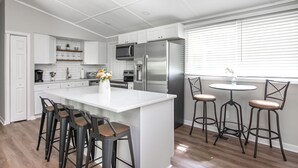 A modern kitchen with white cabinets, a stainless steel refrigerator, a central island with stools, and a window with blinds.