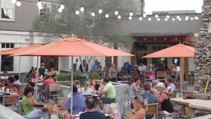 An outdoor dining area with tables and orange umbrellas, surrounded by brick walls and string lights.