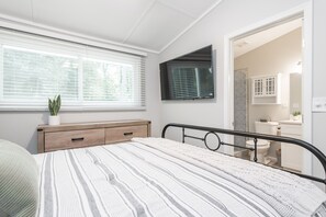 A bedroom with a bed, a bedside table, a window with blinds, a potted plant, and a bathroom visible through an open door.