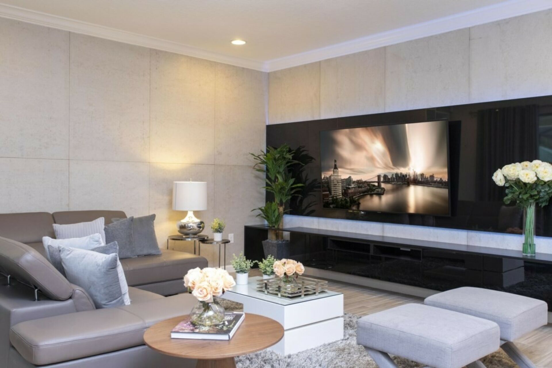 Ein modernes Wohnzimmer mit einem großen Flachbildfernseher, einer Couch, einem Couchtisch mit einem Buch und Blumen sowie einem kleinen Beistelltisch mit einer Lampe.