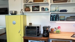 Eine kompakte Küche mit gelbem Kühlschrank, einem Toasterbackofen, einer Mikrowelle und einer Kaffeemaschine. Darüber befinden sich Regale mit verschiedenen Küchenutensilien und einem kleinen Schiffmodell.
