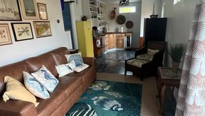 Ein Wohnzimmer mit braunem Ledersofa, einem gemusterten Teppich, einem Deckenventilator und einer Küche im Hintergrund.