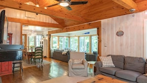 A spacious living room with a wooden ceiling, a ceiling fan, and large windows offering a view of the outdoors. - 5 Bed Cottage & 2 Bunkies! Boathouse Rooftop Deck with Sunsets! (Muskoka Lakes)