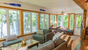 A cozy cabin interior with large windows, wooden beams, a dining area, and a fireplace. - 5 Bed Cottage & 2 Bunkies! Boathouse Rooftop Deck with Sunsets! (Muskoka Lakes)