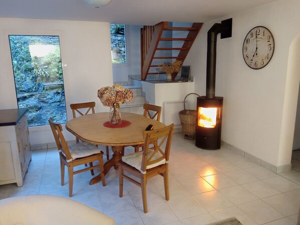 A dining area with a round table, wooden chairs, a vase with flowers, a clock, and a wood-burning stove.