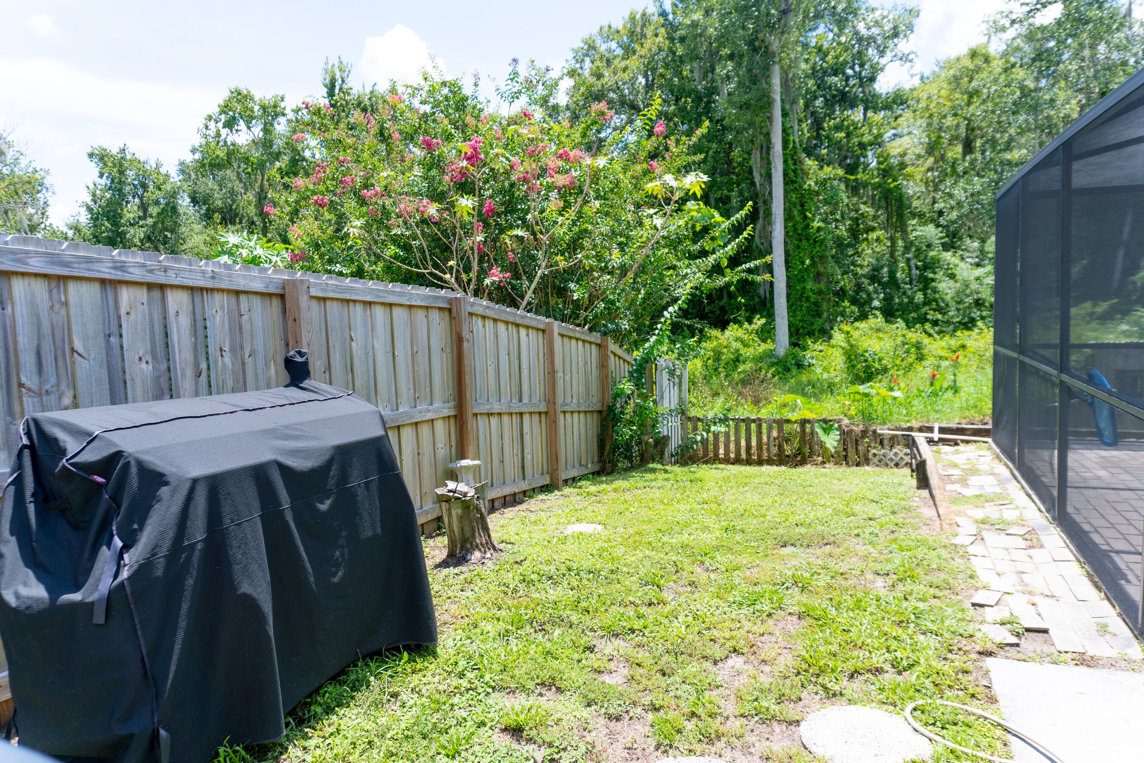A backyard with a covered grill, a wooden fence, and a garden area with trees and plants.