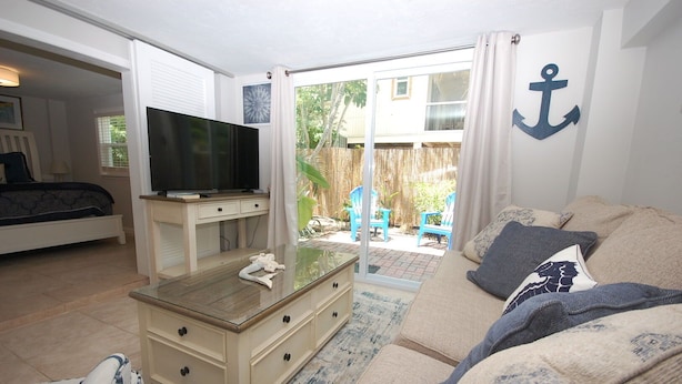 Una sala de estar acogedora con un televisor sobre un mueble de madera, un sofá con cojines estampados y un gran ventanal corredizo que da a un patio trasero con mobiliario exterior.
