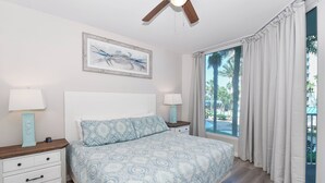 Una camera da letto con un letto, due comodini, una ventola a soffitto, una finestra con tende e un quadro appeso al muro.