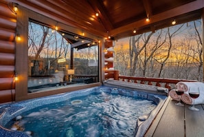 A hot tub with a view of trees and a sunset, wooden deck, and a glass-enclosed area with a lamp.