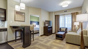 A hotel room with a kitchenette, a dining area, a living room with a sofa, a TV, and a window with curtains. - Great for Business Travelers! Comfortable Suite in St. Louis | BBQ Area (Maryland Heights)