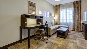 Une chambre d’hôtel équipée d’un bureau, d’une chaise, d’un canapé et d’une petite table. Deux tableaux encadrés ornent le mur, ainsi qu’une lampe et une fenêtre avec des rideaux.