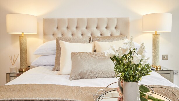 Een opgemaakt bed met een gestuufd hoofdeinde, witte beddengoed en decoratieve kussens. Twee bedlampen met lampenkappen en een vaas met bloemen op het nachtkastje.