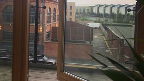 Uitzicht vanuit een raam met houten kader, uitkijkend op een parkeerplaats en gebouwen met bakstenen gevels en moderne constructies.