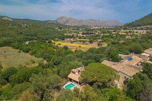 Vue aérienne d’une zone rurale avec une maison dotée d’une piscine, entourée d’une végétation luxuriante et avec des montagnes en arrière-plan.