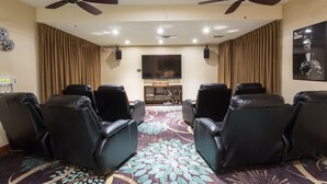 A room with four black leather reclining chairs arranged in a semi-circle, a flat-screen TV on the wall, ceiling fans, and a patterned carpet.