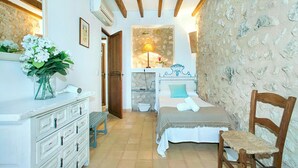 A bedroom with a bed, a wooden chair, a dresser, a lamp, and a stone wall.