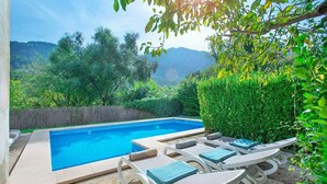 A rectangular swimming pool with a clear blue water surface, surrounded by a low stone wall. There are several lounge chairs with blue cushions arranged around the pool, and a neatly trimmed hedge to the right.