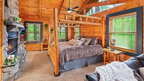Un dormitorio de cabaña de troncos con chimenea de piedra, una cama con manta mullida, un banco de madera y una ventana con persianas.