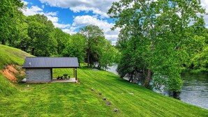 A stone building with a covered patio and picnic tables, situated on a grassy slope overlooking a river.